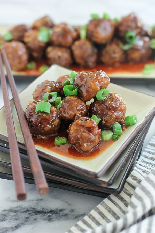 Asian Sweet and sour Meatballs plated on a square plate with chopsticks, with the serving dish with meatballs in the background