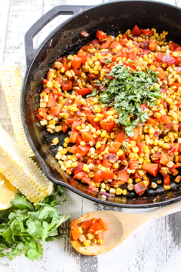 Fiesta corn saute in a cast iron skillet topped with cilantro and a spoon next to the skillet with the corn sauce on it as well as the corn cobs with the corn sliced off